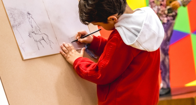 Een jongen in een rode trui tekent op een ezel, met een kleurrijke achtergrond.