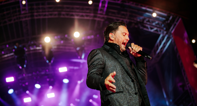 Ein Sänger in einem glänzenden Anzug singt leidenschaftlich auf einer Bühne mit bunten Lichtern.