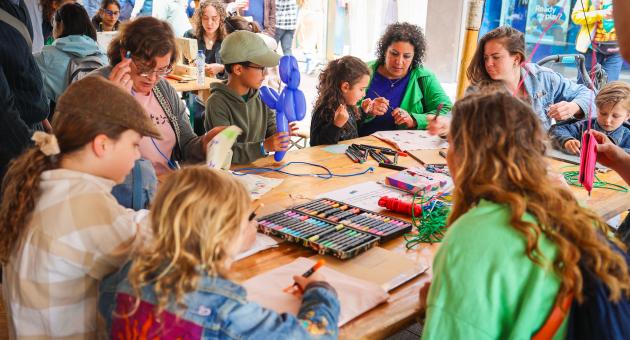 Groep kinderen aan een grote tafel vol knutselspullen die een workshop doen onder begeleiding van twee volwassen vrouwen.