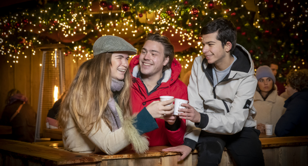 Een meisje en twee jongens proosten met warme chocomel in een sfeervol verlichte houten kraam tijdens de Royal Christmas Fair op Lange Voorhout