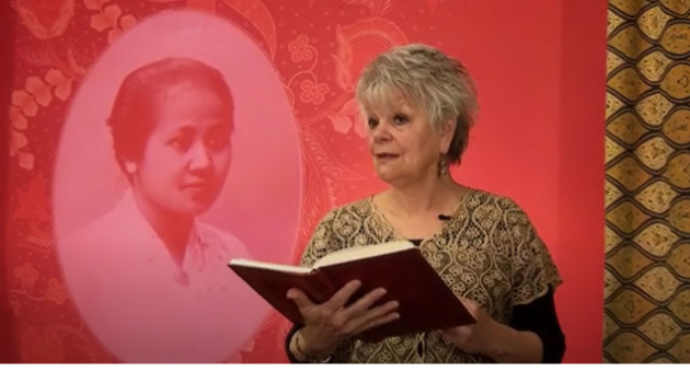 Anita Poolman draagt voor met een boek in haar hand, staand voor een portret van Kartini; dochter van de regent van Japara
