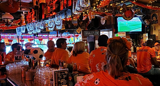 Mensen in oranje shirts kijken naar een wedstrijd in een bar, met versiering en een tv.