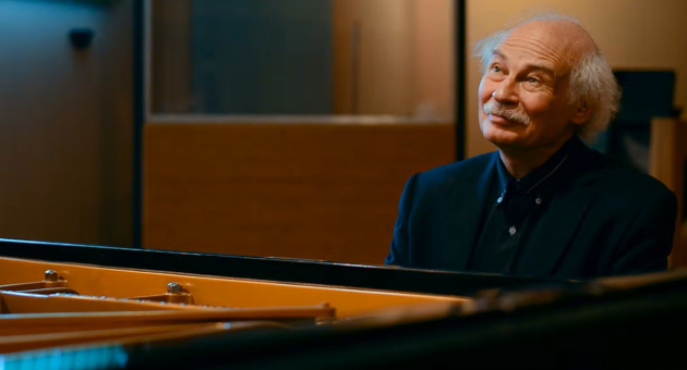 Een man met grijs haar zit met een glimlach achter een piano.