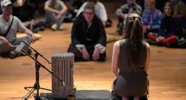 A woman performs for a group of people, with a musical instrument in front of her.