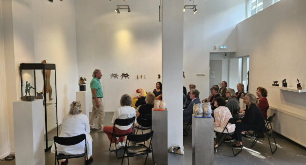 Groep mensen zit op klapstoelen in een witte galerie, luistert naar spreker tussen keramische kunstobjecten