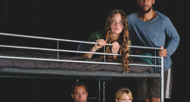 Vier mensen rondom een metalen stapelbed op een toneel, met zwarte achtergrond en serieuze blikken