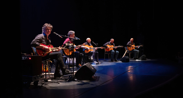 Vijf muzikanten zitten in een rij op een donker podium en spelen akoestische gitaren onder spots
