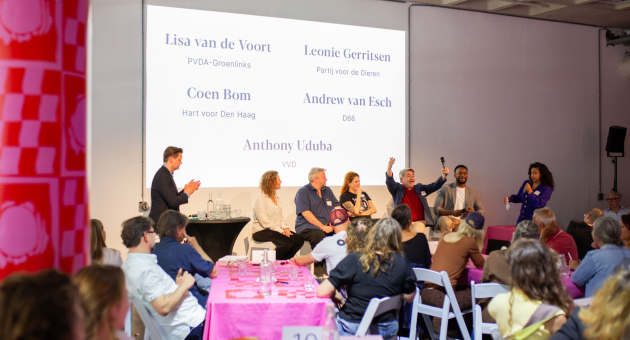 Een panel haagse politici zit op een podium. In de zaal zit publiek. 