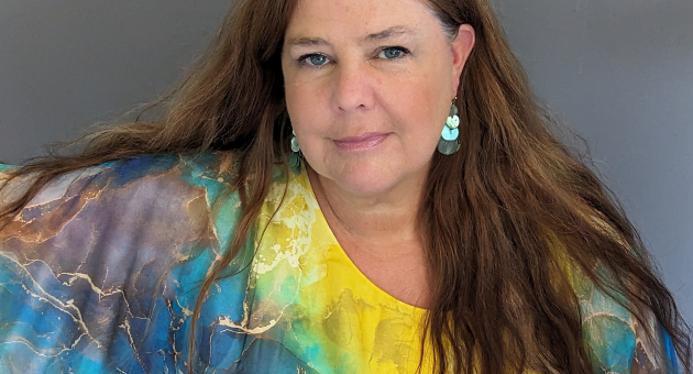 Vrouw met lang bruin haar en blauw-geel blouse poseert.