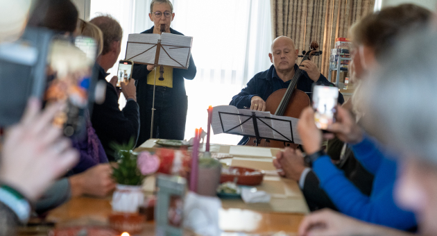 Musicians play the recorder and cello for a group of people seated at a long table in a living room.