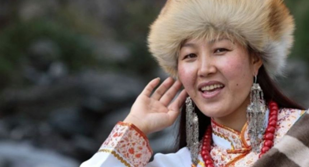 Een vrouw met een bonthoed en traditionele kleding, glimlacht en steekt haar hand op.