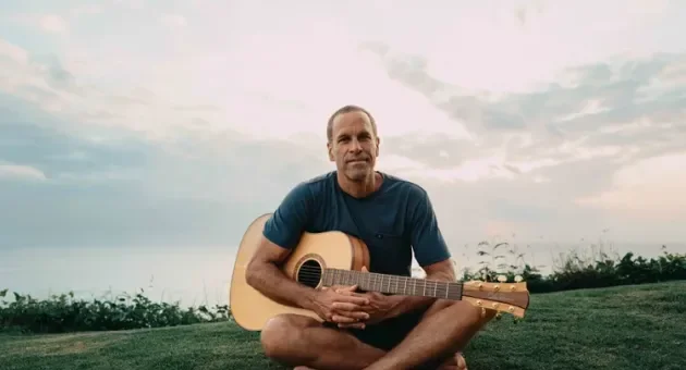Man zit op het gras met een gitaar, kijkend naar de horizon.