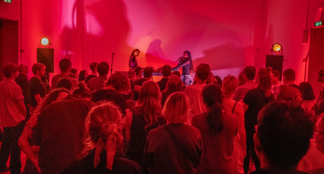 Groep mensen op de rug gezien die in een rood verlichte zaal naar twee muzikanten op een podium kijken.