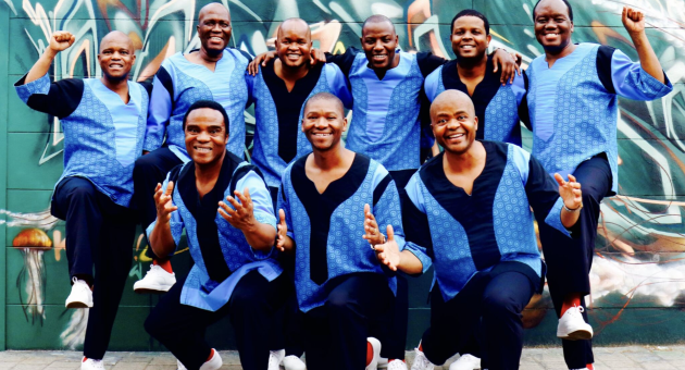 Negen mannen in traditie-inspirerende kleding met blauwe tinten poseren vrolijk voor een muurschildering.