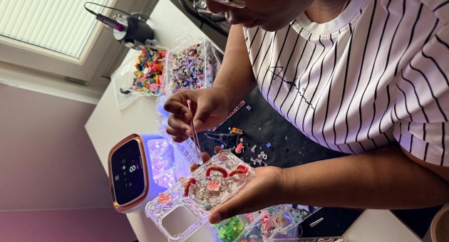 Persoon versiert een doorzichtige telefoonhoes met kralen en figuurtjes aan een bureau vol knutselspullen