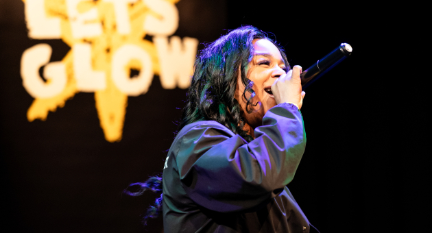 Zangeres met microfoon op podium voor zwart decor met tekst ‘LET’S GLOW’ in fel verlichte letters