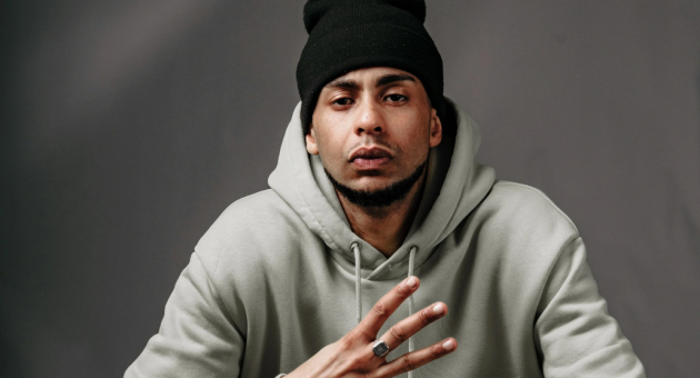 Man met zwarte muts en grijze hoodie maakt handgebaar met vier vingers tegen neutrale achtergrond