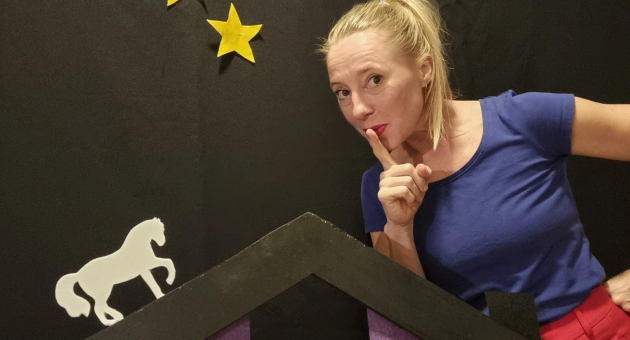 Vrouw in blauwe top legt vinger op haar lip, naast decor met sterren en wit paard op zwart doek