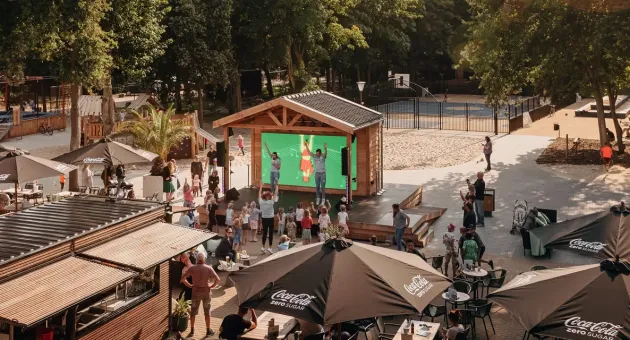 Een drukke recreatieplek met mensen, parasols en een scherm.