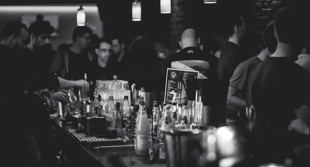 Drukke bar met mensen aan de toog, flessen drank en glazen op de voorgrond, sfeerverlichting boven de bar