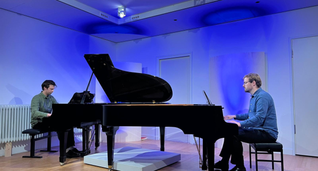 Twee pianisten spelen op vleugels tegenover elkaar in een blauwe verlichte zaal met houten vloer