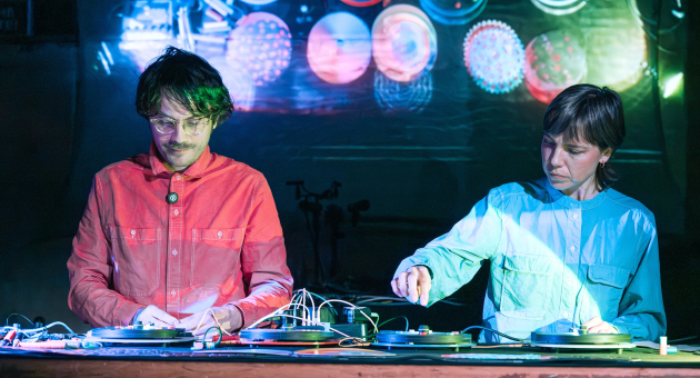 Twee dj’s in felgekleurde outfits mixen live op draaitafels met visuele projecties op de achtergrond