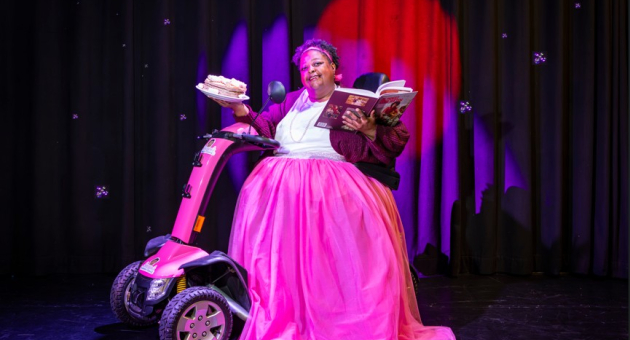 Persoon in roze jurk op roze scootmobiel met taart en boeken op het podium voor een zwart gordijn met paarse spotlights