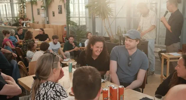 Groep mensen zit aan tafel in een lichte ruimte met planten, lachen en praten.