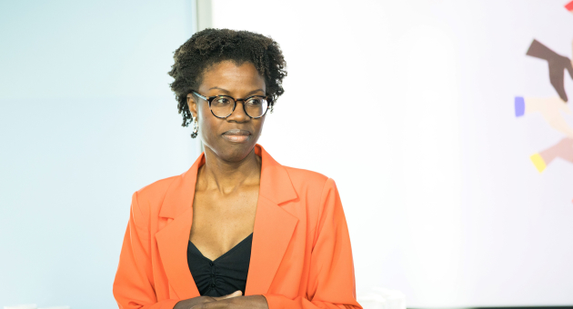 Een vrouw in een oranje blazer spreekt met een serieuze houding.