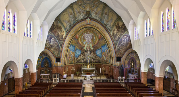 Interieur van een kerk met houten banken, kleurrijke mozaïeken en een groot altaar in het midden