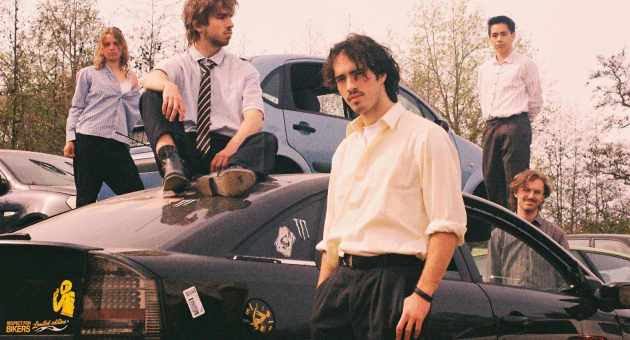Vijf jongeren van band See Sharp staan bij een zwarte auto op een parkeerplaats.