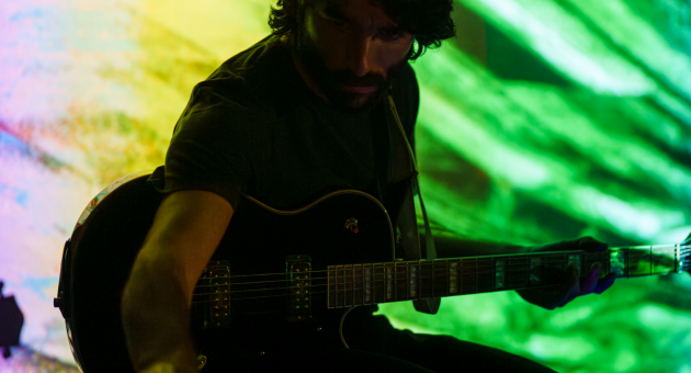 Man met elektrische gitaar op een podium met felgekleurde, abstracte achtergrondverlichting