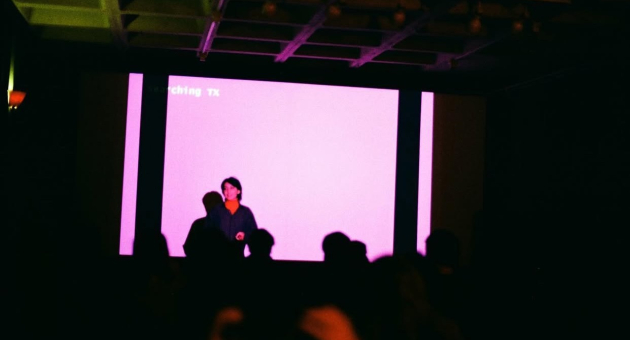 Persoon presenteert voor roze projectiescherm in donkere ruimte met publiek op de voorgrond