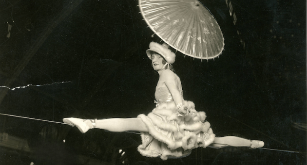 Een acrobate in een witte jurk met een parasol, die op een koord balanceert.