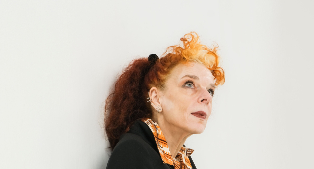 Een vrouw met een oranje haar en een zwart jasje zit tegen de muur.