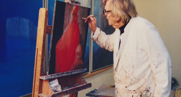 Kunstenaar in witte jas schildert op een ezel in een atelier, omringd door blauwe doeken aan de muur