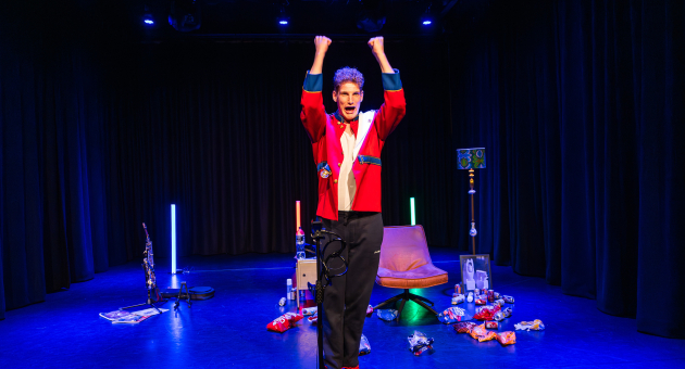 Een performer in een rode jas staat met opgestoken armen op het podium.