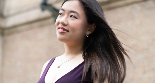 Vrouw met lange haren en een paarse jurk, staand bij een muur.