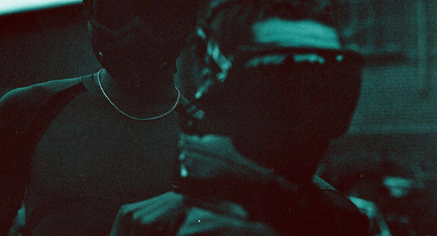 Twee mensen met donkere maskers en kappen in de schaduw.