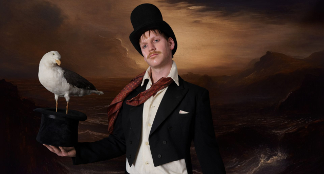Man in zwart kostuum met hoge hoed houdt een meeuw op een tweede hoed, met dramatisch zeegezicht op de achtergrond