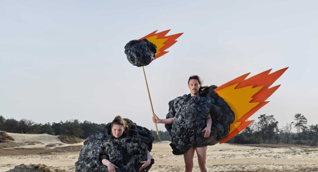 Twee personen verkleed als vurige meteorieten in zanderig duinlandschap met blauwe lucht