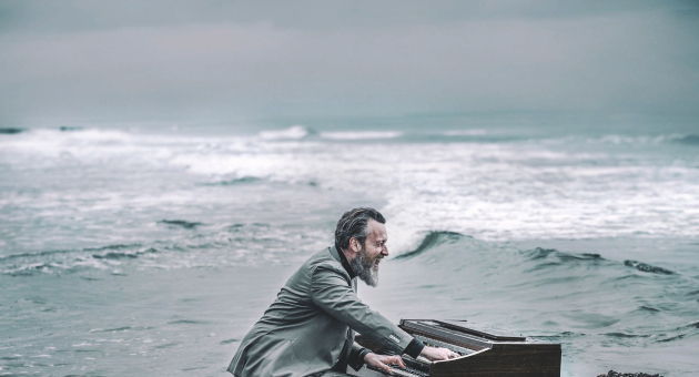 Man in grijs pak speelt piano in ondiep zeewater bij een ruige, bewolkte kustlijn.