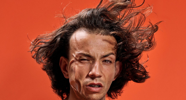 Portret van een man met wapperend haar en verwrongen gezichtsuitdrukking, tegen een feloranje achtergrond.