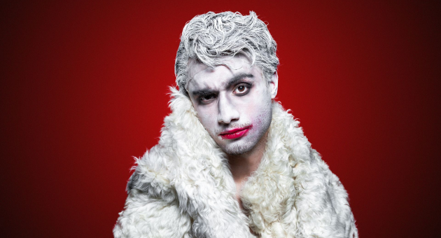 Persoon met wit geschminkt gezicht en haar, rode lippenstift en witte bontjas, poseert voor een rode achtergrond.