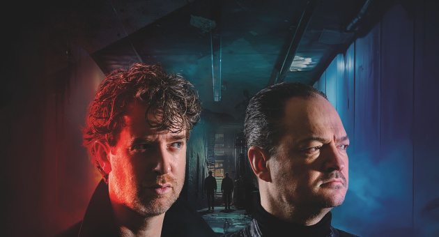 Twee mannen met serieuze gezichten staan in een donker, mysterieus verlicht gangpad; de ene belicht met rood licht, de andere met blauw.