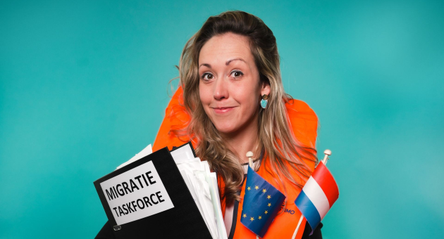 Close up van Djûke Stammeshaus poserend voor een blauwe achtergrond. Ze draagt een oranje reddingsvest, een Europese en Nederlandse vlag en een uitpuilende map met migratie taskforce erop. 