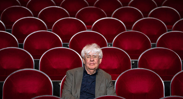 Oudere man met grijs haar zit alleen op een rij in een lege zaal met rode theaterstoelen