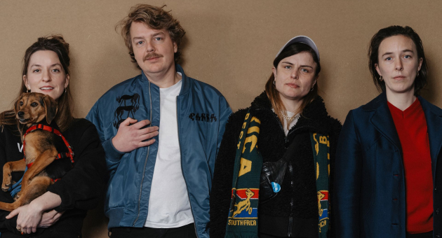 Vier mensen poseren voor beige achtergrond, één met een hondje in de armen