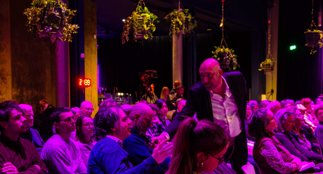 Een presentator in pak geeft een man uit het publiek een microfoon aan. Het publiek zit op stoelen in een roze verlichte zaal 