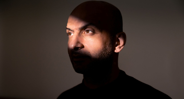 Man met kaal hoofd in donkere ruimte, gedeeltelijk verlicht door een diagonale lichtstreep over zijn gezicht.